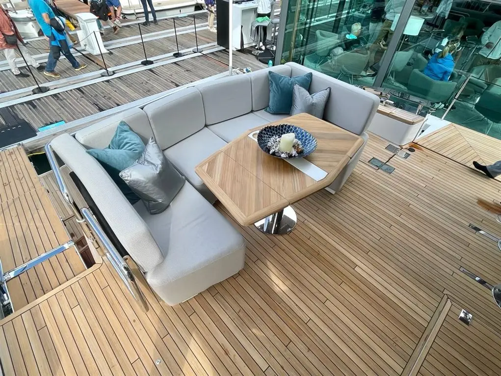 Modern yacht deck featuring a stylish seating area with grey cushions and a wooden table, surrounded by people and a marina.